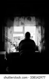 Grandfather Looks Out Window. Silhouette Of Old Man In Dark Room. Lifestyle In Old Age. Loneliness, Sad Mood. Black And White Photo