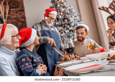 Grandfather in santa hat carving chicken at christmas dinner at home. Active seniors concept. New Year Eve celebration. Festive table with family reunion - Powered by Shutterstock