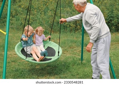 Grandfather swinging children in summer park. Grand dad and grandchildren sitting on swing outdoors. Senior 60s Grandpa pushing small grandkids on a rope seesaw. Old man and little kids at playground