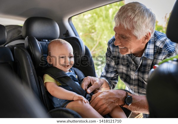 Grandfather tying baby in child seat. Cute little\
boy going for road trip with grandfather. Happy senior man and\
happpy smiling grandchild enjoying car trip. Toddler boy buckled\
into car seat.