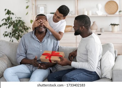 Grandfather's Birthday. Mature Black Man And His Preteen Son Congratulating Grandad, Surprising Him With Gift Sitting On Couch At Home