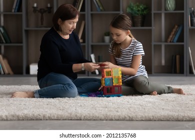 Grandkid And Grandma Playing At Home, Engaged In Building Game On Heating Floor With Carpet In Living Room, Constructing Block Tower. Older Nanny Watching Child. Family, Intergenerational Relationship