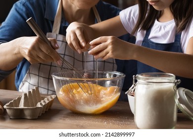 Grandkid helping grandma to cook omelet for breakfast, cracking beating eggs with whisker in bowl at kitchen table. Grandmother teaching grandchild to bake, preparing dough. Family activity. Close up