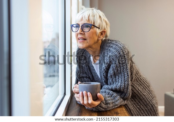 Grandma\
from the 60\'s starts the day with her favorite drink while standing\
and holding a cup of coffee in her hands looks through the windows\
looks satisfied feels good, the older\
generation