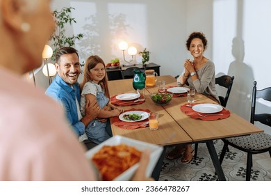 Grandma bringing pasta to the dining table for her family while family is sitting happily and waiting for lunch