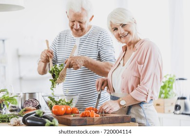 Grandma Cuts Pepper And Grandpa Mixes Vegetable Salad During Cooking Together In White Kitchen