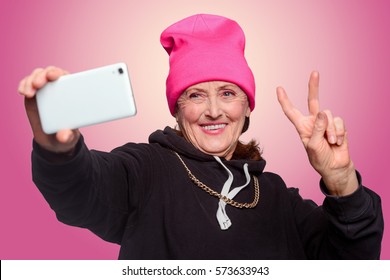 Grandma makes a selfie. Elder woman in black hoodie makes a piece sign and smiles at frontal camera of her smartphone. Dressed beyond her years granny captured in studio shot with pinkish background.