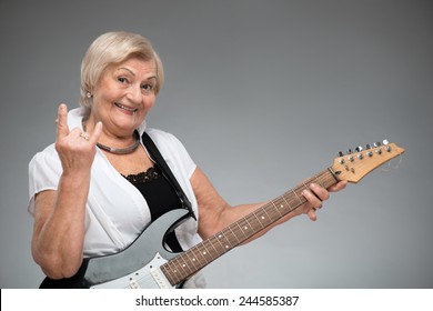 Grandma Playing The Guitar. Closeup Of Funny Smiling Senior Woman Playing The Guitar And Showing Devils Horns While Standing Against Grey Background