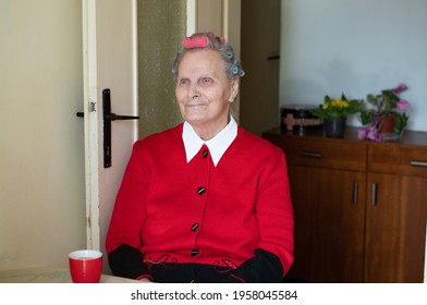 Grandma With A Smile Is Sitting At A Table With Coffee. The Older Woman Has Curlers On Her Head, And Red Blouse. Behind Her A Hallway With Flowers And A Brown Box As Well As A Wooden Chest Of Drawers