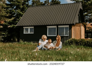 Grandmother with adult daughter and grandson relaxing outdoors in countryside. Happy multi generation family sitting on grass by modern country house, reading book and spending holiday time together. - Powered by Shutterstock