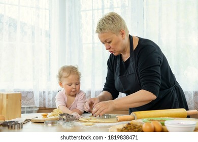 Grandmother In Black Apron Teaches Her Cute Little Granddaughter How To Make Gingerbread Cookies. Little Girl And Grandmother Cooking Together In Kitchen At The Wooden Table. Happy Family Concept.