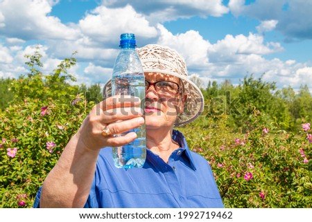 Grandmother with a bottle of water. Elderly woman with a bottle of drinking water on the background of a summer landscape. Hot Summer.
