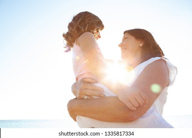 Grandmother Carrying Grand Daughter Enjoying Sunny Beach Holiday Together, Joyful Smiling With Sun Flare Blue Sky, Outdoors. Senior And Child Playful Fun Bonding Family Travel Activities Lifestyle.