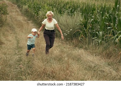 Grandmother And Child Running Outdoor. Happy Family On A Road Near Corn Field During Summer Sunset. Generation, Happiness Vitality Concept.