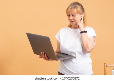 Grandmother In The Digital Age. Closeup Of Thoughtful Senior Woman Trying To Cope With Computer While Standing Against Orange Background. Female In Wireless Headphones And Black Fitness Bracelet Watch