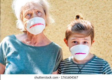 Grandmother With Grandchild In A Respiratory Masks Plays Together Near House. Tired Of Wearing A Mask.Family Fun. Drawing A Sad Smile On Protective Masks. Quarantin, Isolated. Coronavirus Covid-19.