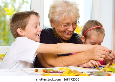 Grandmother With Grandchildren Applying A Dry Maple Leaves Using Glue While Doing Arts And Crafts