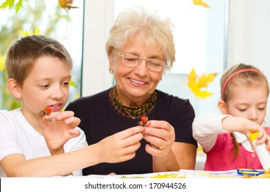 Grandmother With Grandchildren Applying A Dry Maple Leaves Using Glue While Doing Arts And Crafts