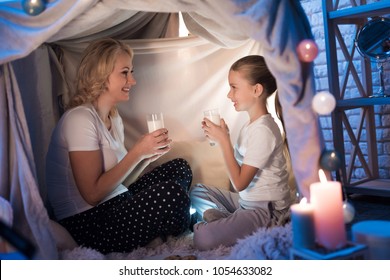 Grandmother and granddaughter are eating cookies with milk in cozy blanket house at night at home.
