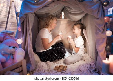 Grandmother And Granddaughter Are Eating Cookies With Milk In Cozy Blanket House At Night At Home.