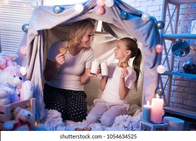 Grandmother And Granddaughter Are Eating Cookies With Milk In Cozy Blanket House At Night At Home.