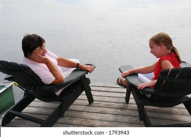 Grandmother And Granddaughter Sitting In Adirondack Chairs On A Dock Having A Conversation