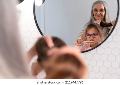Grandmother with granddaughter standing indoors in bathroom, brusing teeth and hair in morning