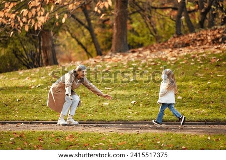 A grandmother greeting her granddaughter, crouching and opening her arms to the sides. Grandmother walking with her granddaughter in the park in autumn.