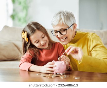 Grandmother and her granddaughter Putting Coin Money In Piggybank  At Home. Personal Savings, Bank Safety And Financial Investments Concept