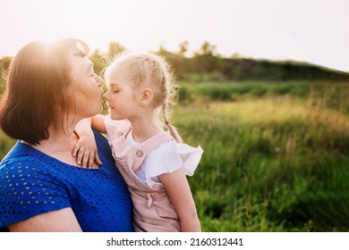 Grandmother holding granddaughter on her arms and kissing nose. Happy childhood, family love concept. Support and togetherness. Two people outdoor with sunlight background. High quality photo