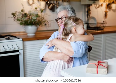 Grandmother Is Hugging Child Girl In Cozy Kitchen At Home. Happy Family Is Giving Gifts And Thanks. Cute Kid And Senior Woman Is Enjoying Kindness. Lifestyle Moment. Holidays Hanukkah, Thanksgiving.