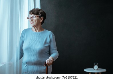 Grandmother With Insomnia Holding Cane In Room With Clock On Table Against Black Background