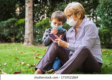 Grandmother and little kid playing with a smartphone in the backyard while wearing protective masks due to coronavirus outbreak