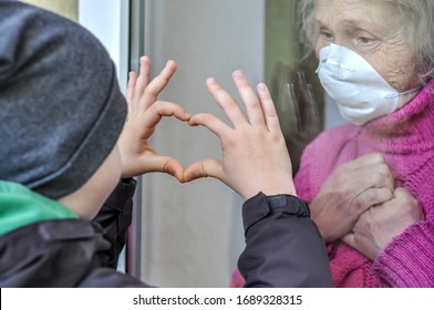 Grandmother Mature Woman In A Respiratory Mask Communicates With Her Grandchild Through A Window. Elderly Quarantined, Isolated. Coronavirus Covid-19. Caring With Older People. Family Values, Love