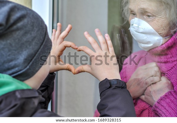 Grandmother mature woman in a respiratory mask\
communicates with her grandchild through a window. Elderly\
quarantined, isolated. Coronavirus covid-19. Caring with older\
people. Family values,\
love