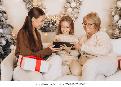 A grandmother, mother, and daughter share a joyful moment on the couch surrounded by Christmas decorations and gifts. They engage with a digital tablet, embodying the festive holiday spirit. - Powered by Shutterstock