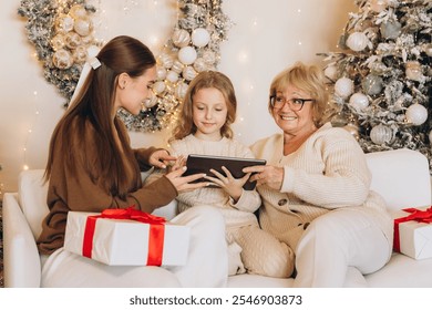 A grandmother, mother, and daughter share a joyful moment on the couch surrounded by Christmas decorations and gifts. They engage with a digital tablet, embodying the festive holiday spirit. - Powered by Shutterstock