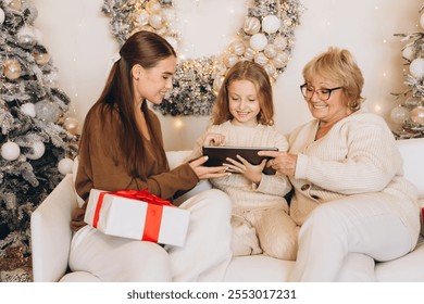 A grandmother, mother, and daughter share a joyful moment on the couch surrounded by Christmas decorations and gifts. They engage with a digital tablet, embodying the festive holiday spirit. - Powered by Shutterstock