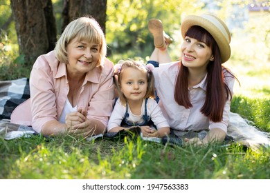 Grandmother, Mother And Grand Daughter Enjoying Sunny Garden Holiday Together Outdoors, Lying On Green Grass On Blanket And Smiling To Camera. Leisure Family Lifestyle, Happiness And Moments.