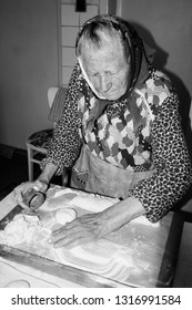 A Grandmother, Old Woman, Senior Woman Is Baking, Cooking In The Kitchen. MiddleEuropean Woman Is Wearing A Traditional Clothes. Middleeuropean, Central Culture.Black And White Portrait. Retro,vintage