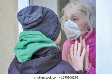 Grandmother In A Respiratory Mask Communicates With Her Grandson Through A Window. Elderly Quarantined, Isolated. Pandemic Coronavirus Covid-19. Caring With Older People. Family Values