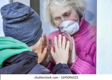 Grandmother In A Respiratory Mask Plays With Hands With Her Grandson Through A Window. Elderly Quarantined, Isolated. Pandemic Coronavirus Covid-19. Caring With Older People. Family Values