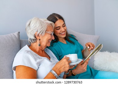 Grandmother Sitting On A Couch In A Sunny Room, Looking At A Photo Album And Sharing Fond Memories With A Tender Caregiver. Young Caregiver And Senior Woman Laughing Together While Sitting On Sofa 