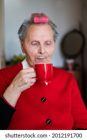 Grandmother With A Smile Is Sitting At A Table And Drinking A Coffee. The Older Woman Has Curlers On Her Head, And Red Blouse. Enjoying Morning In Her Apartment. Portrait Of Old Women
