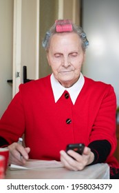 A Grandmother With A Smiling Face Looks At Her Cell Phone. The Older Woman Has Pink And Blue Curlers On Her Head. She Is Wearing A White Shirt Over Which Is A Red Sweater.