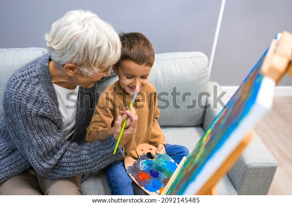 Grandmother spending happy\
time with grandson. Woman assisting young boy in painting on\
canvas. Happy senior woman looking at her grandson painting and\
smiling at him