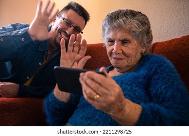 Grandmother Talk On Webcam Phone Call With Her Grandson. Grandma In Blue Sweater Sit On Sofa For Video Conference With Smartphone. Old Caucasian 90s Woman Feel Happy On Couch At Living Room Home.