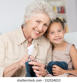 Grandmother Teaching Her Pretty Smiling Little Granddaughter To Knit As They Sit Close Together On A Sofa In The Living Room