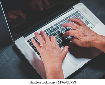 Grandmother Works Behind Laptop, Typing On Keyboard With Her Old Hands With Wrinkles. Modern Old Pensioner Working From Home. Close Up View Of Elderly Woman's Hands