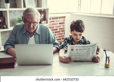 Grandpa And Grandson Resting At Home. Handsome Old Man Is Using A Laptop While His Boy Is Reading A Newspaper